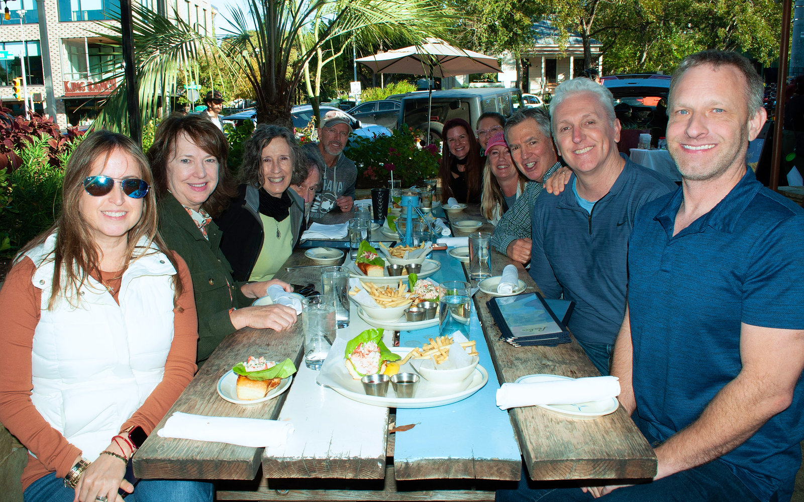 A group of guests smiling around a shared outdoor table of lobster rolls and fries
